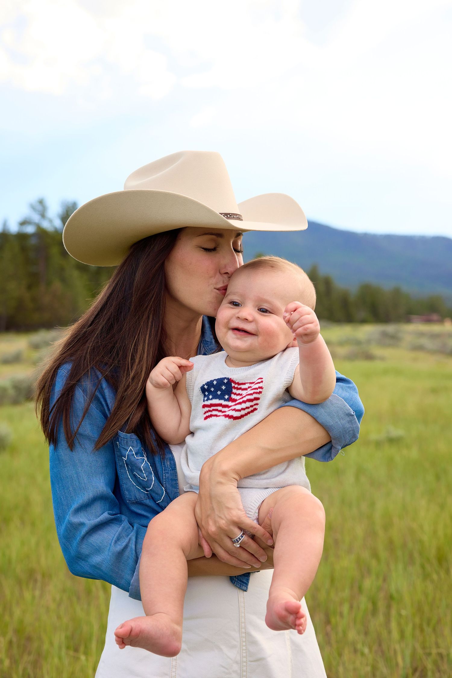 A woman in a cowboy hat kisses a happy baby wearing an American flag onesie outdoors.