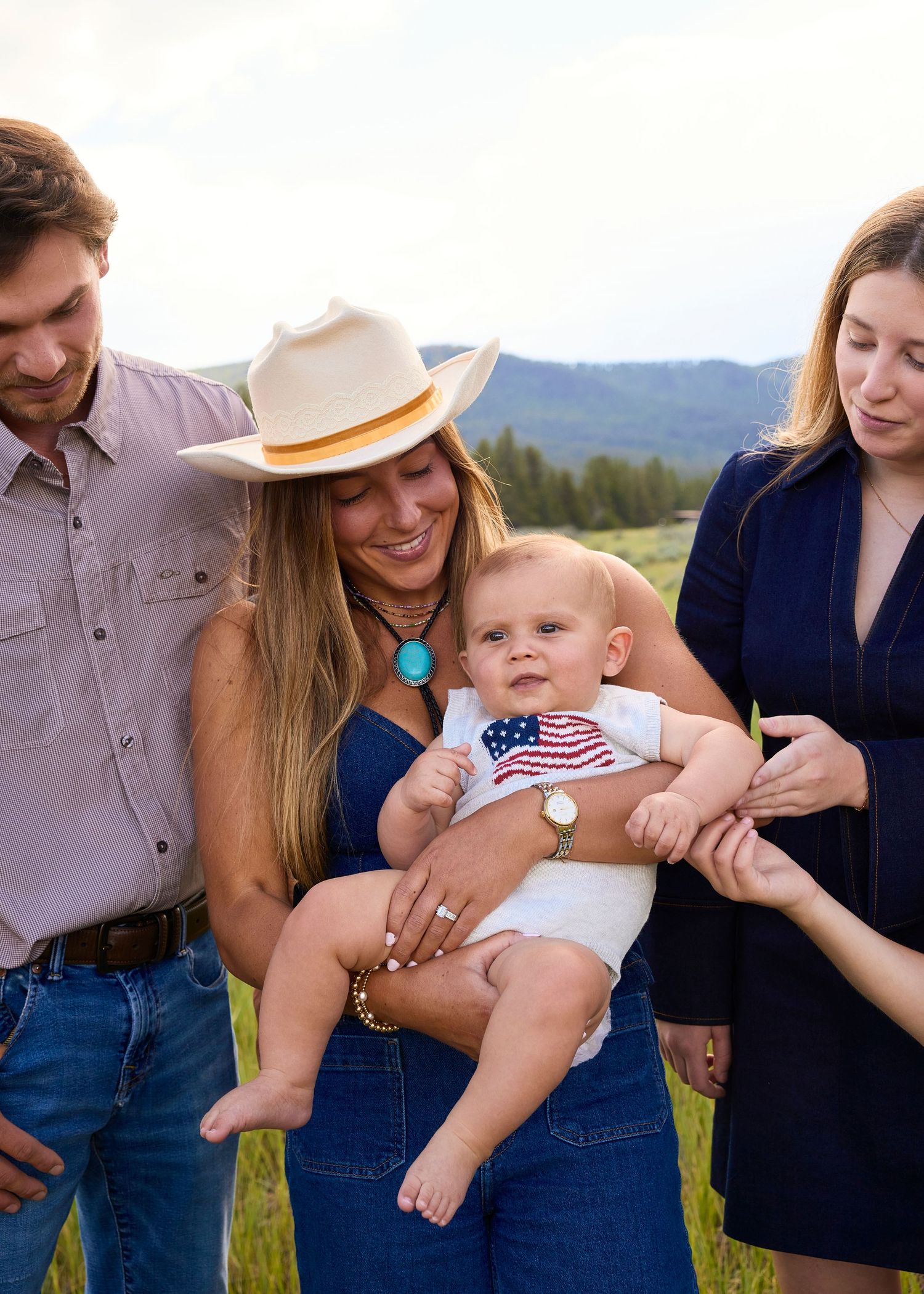 A smiling woman in a cowboy hat holds a baby in an American flag onesie outdoors.