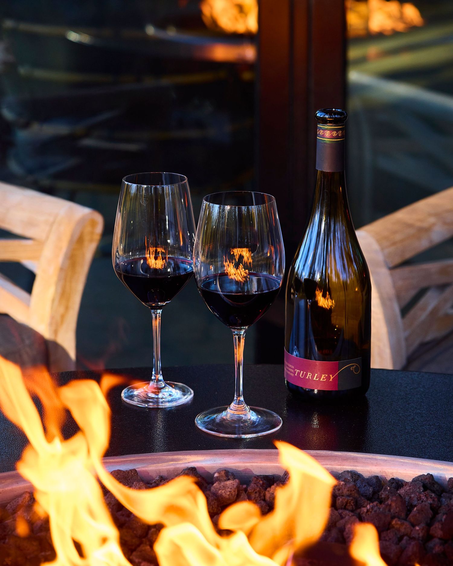 Two glasses of red wine and a bottle sit beside a blazing outdoor fire pit.