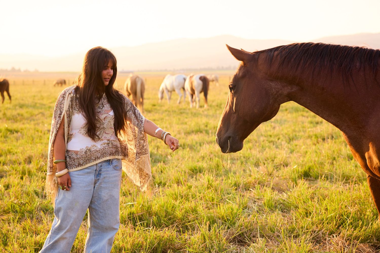 Boho-style woman interacting with a chestnut horse in a glowing sunset field with grazing horses.