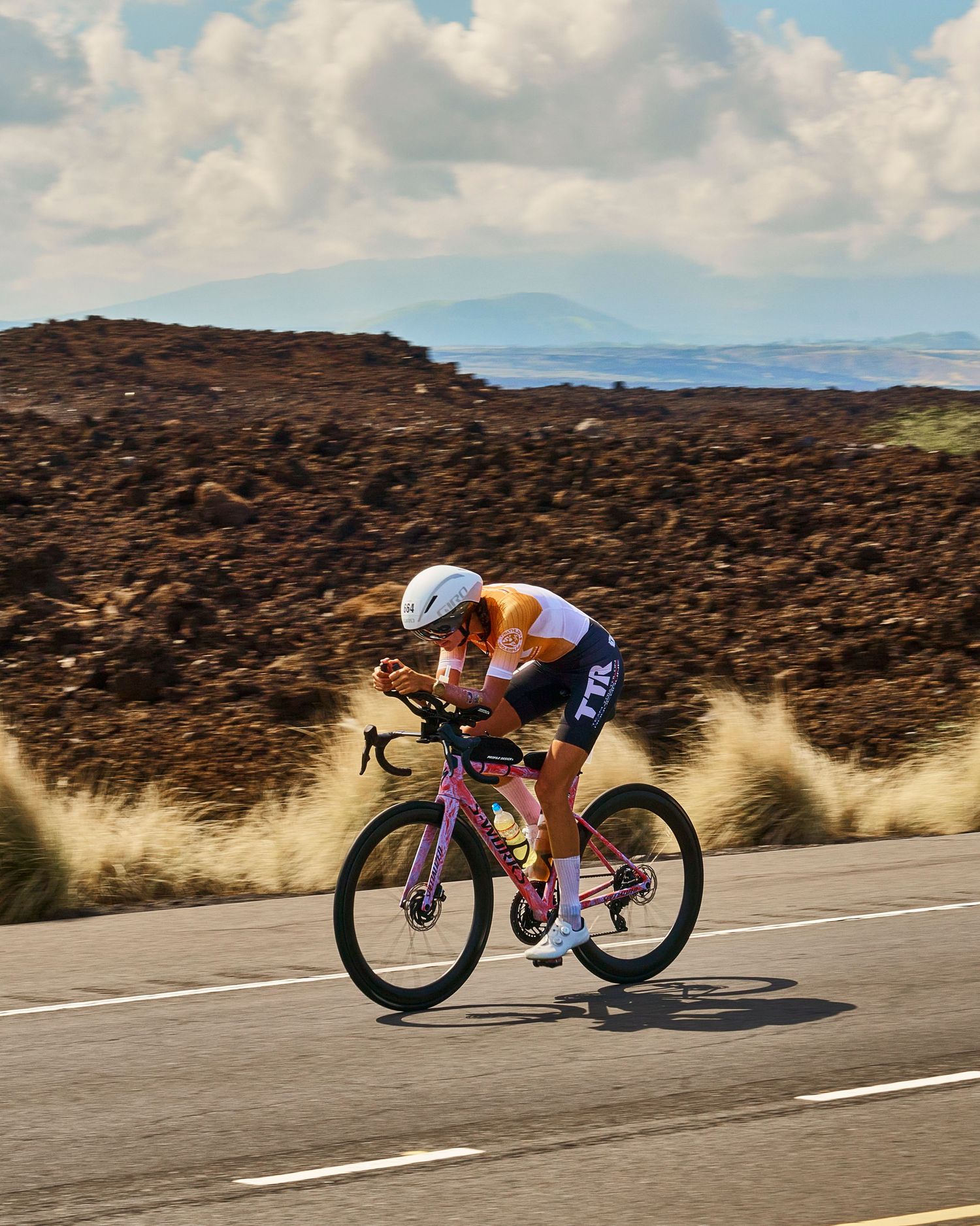 Triathlete in aerodynamic position rides a pink bike on a road during an Ironman race with volcanic landscape behind.