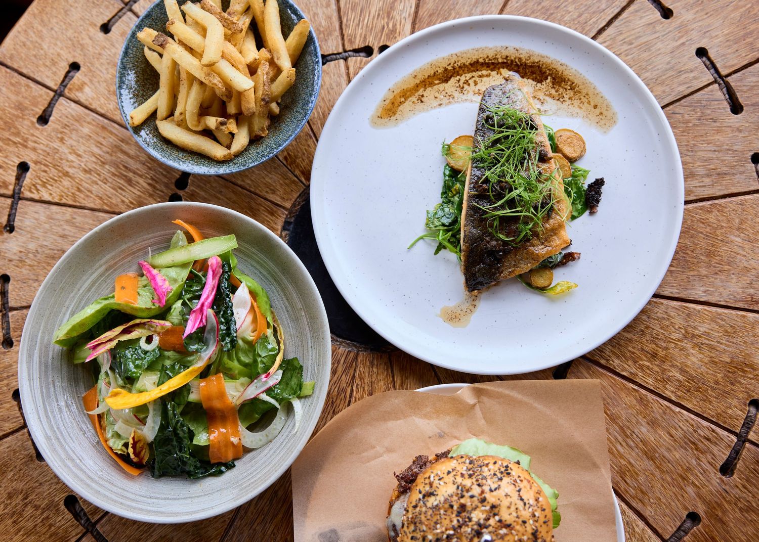 Overhead view of gourmet dishes including grilled fish, fresh salad, fries, and a burger on a wooden table.