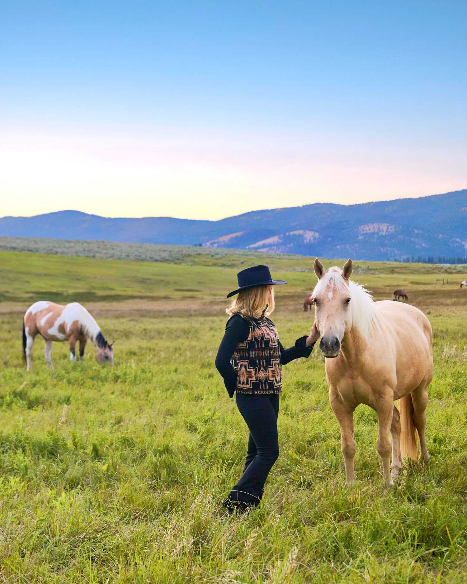 Woman in black hat petting palomino horse in green mountain pasture at sunset.