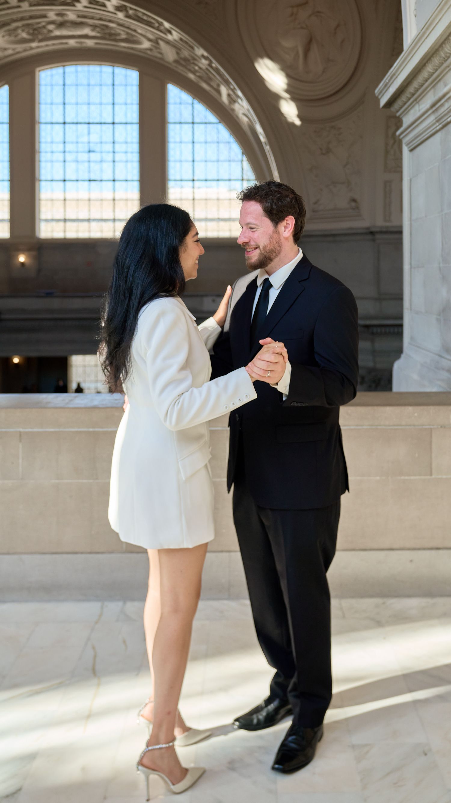Newlyweds dancing together in sunlit San Francisco City Hall rotunda, bride in white blazer dress and groom in black suit.