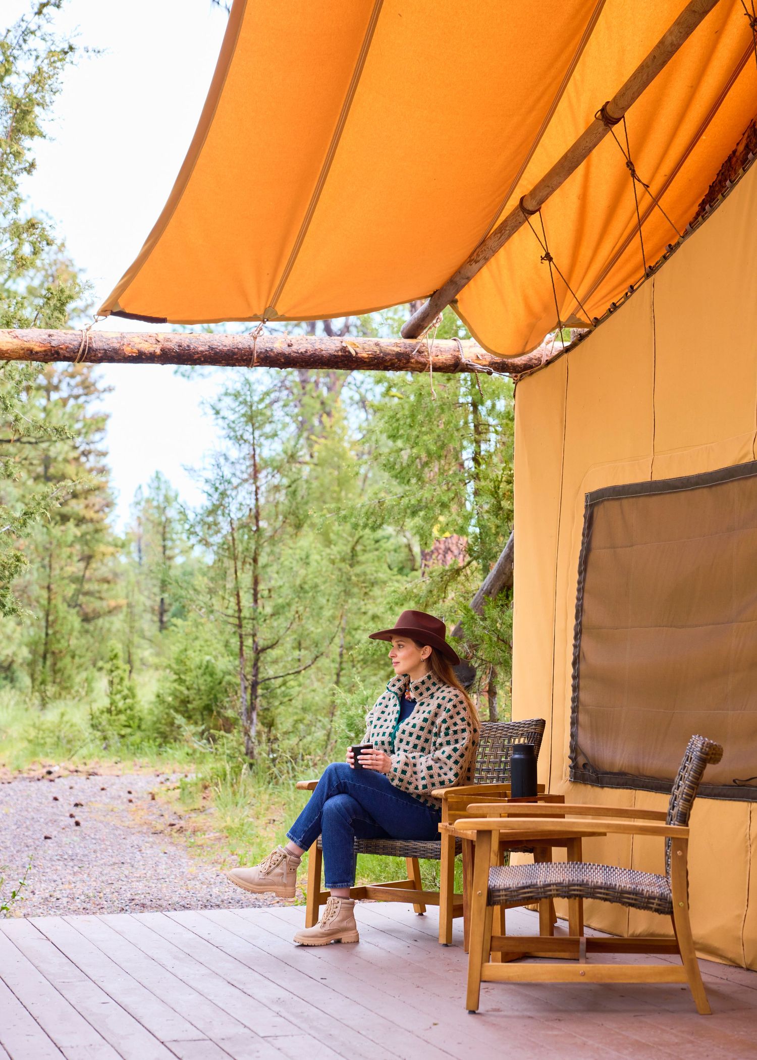 Woman in cowboy hat relaxes on a glamping tent deck surrounded by lush forest trees.