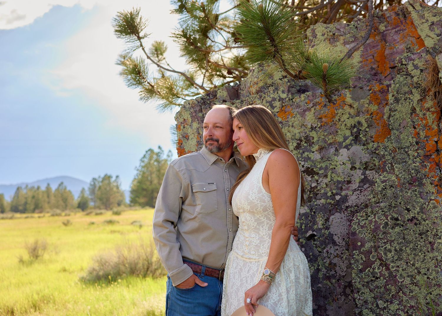 Couple leaning against a lichen-covered rock wall in a mountain meadow, sharing a tender romantic moment outdoors.