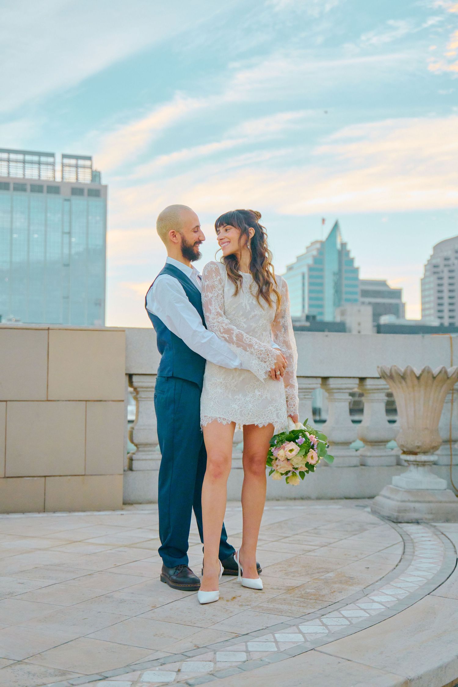 Bride in short lace dress holds bouquet while groom in blue vest embraces her on rooftop with city skyline.