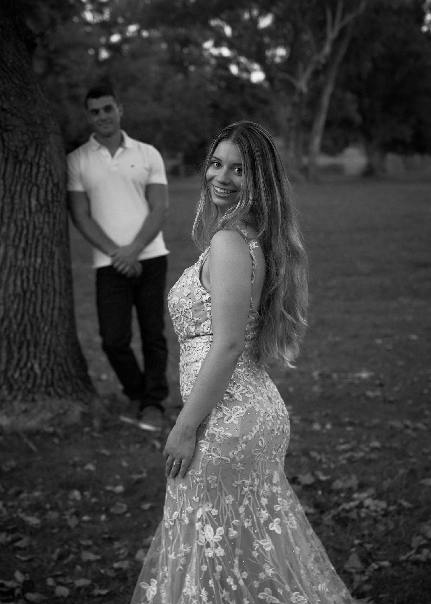 Black and white photo of bride in lace gown smiling with groom in background by tree.