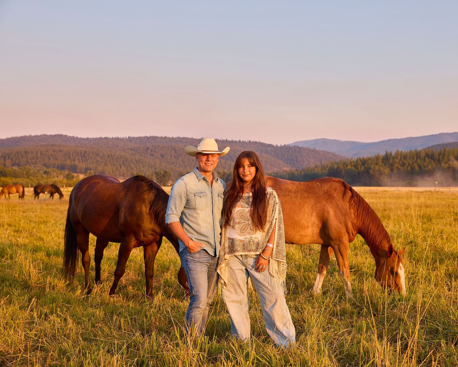 Couple standing together in field with grazing horses and mountain backdrop at golden hour.
