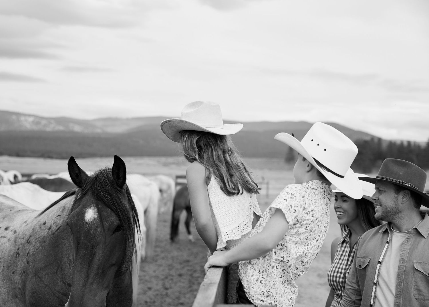 Two women in cowboy hats petting a horse at a ranch with mountains in the background, black and white.