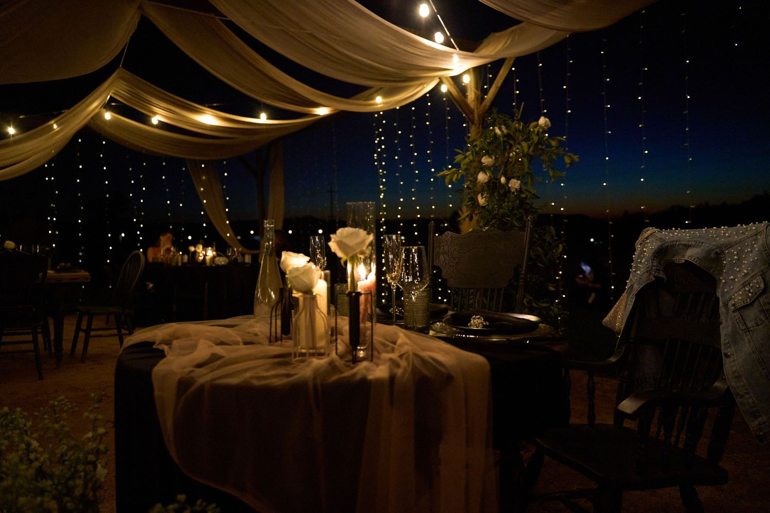 Romantic nighttime wedding reception under draped canopy with string lights, candles, and floral centerpieces on rustic tables.