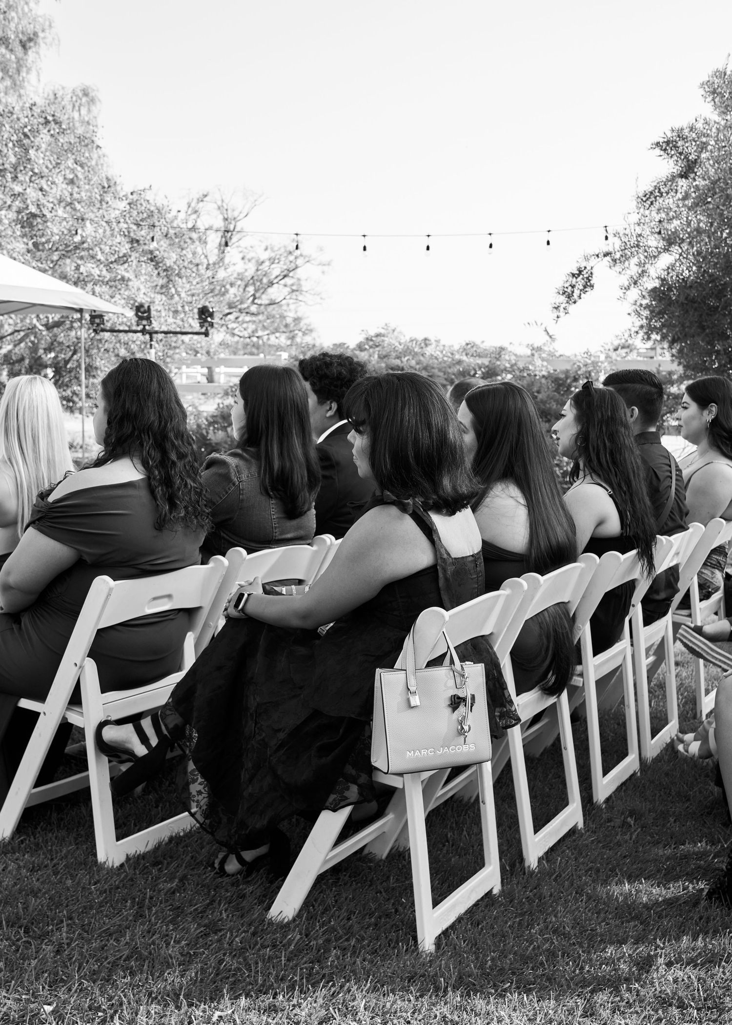 Black and white photo of wedding guests seated in white folding chairs at an outdoor ceremony.