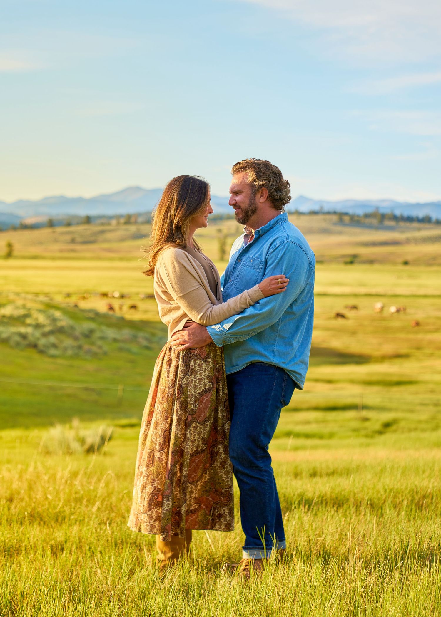 Couple embraces in golden meadow at sunset with rolling hills and mountains in background during engagement shoot.