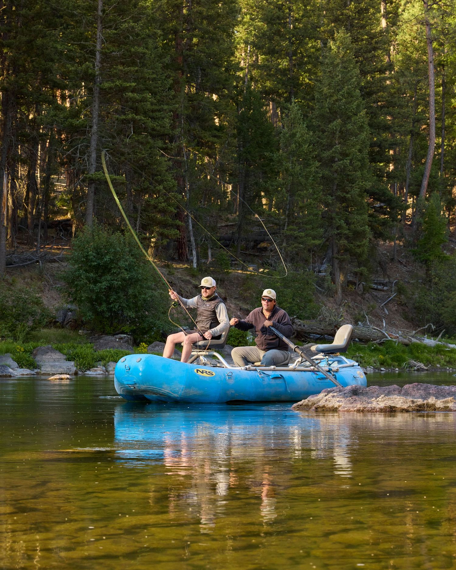 Two anglers fly fishing from a blue inflatable raft on a calm mountain river surrounded by pine forest.