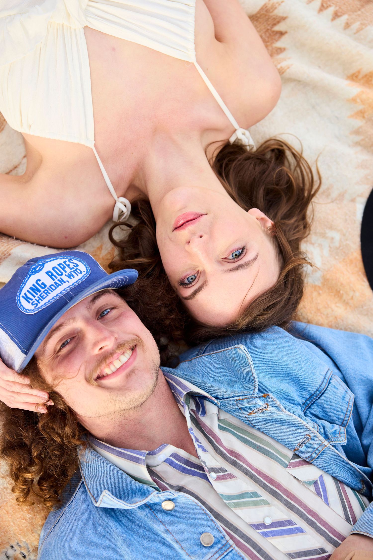 Smiling couple lies on blanket looking up at camera, man in denim jacket and blue trucker hat.