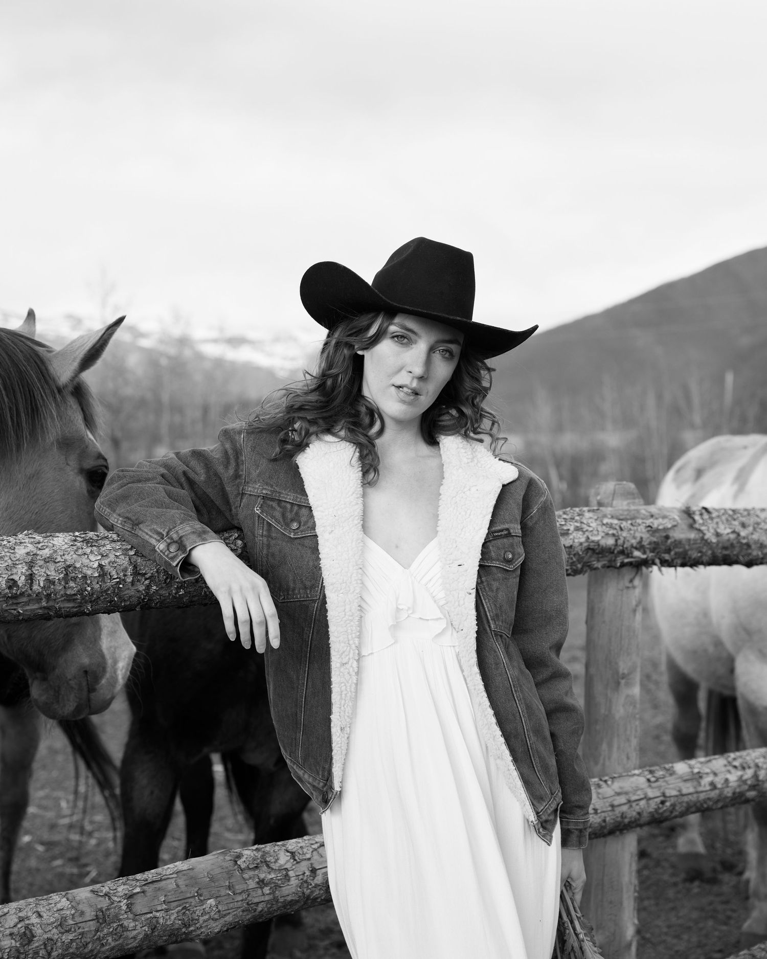 Woman in black cowboy hat and denim jacket posing with horses at a rustic fence.