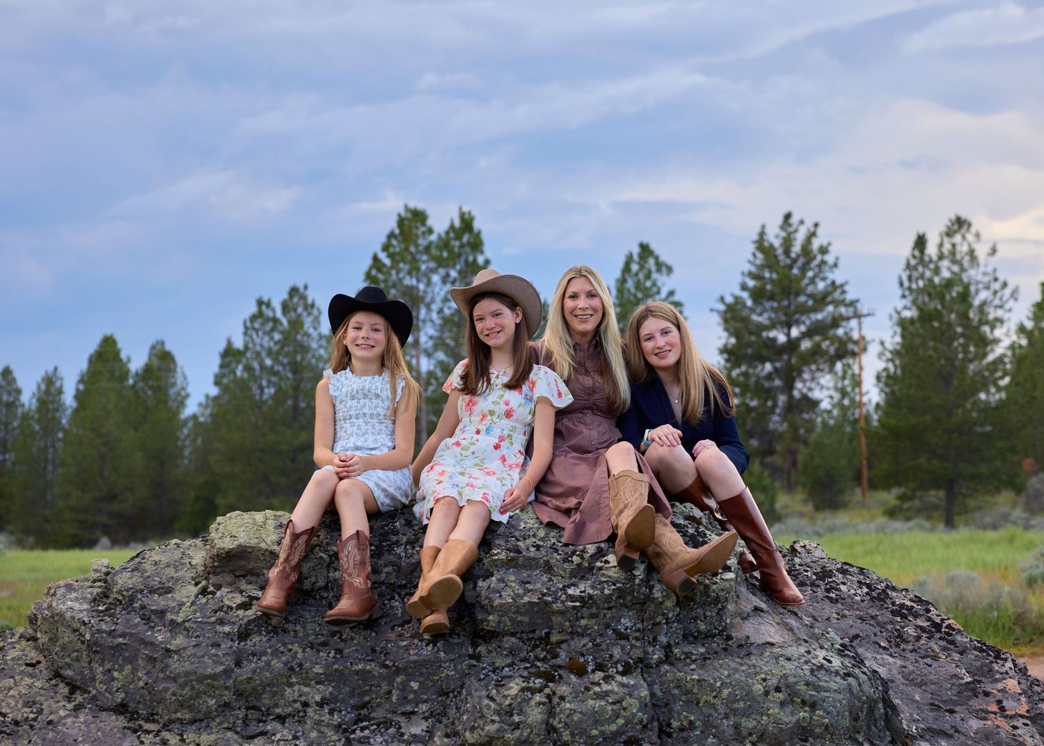 Four girls sitting on a large rock in cowboy boots and hats, smiling against a pine tree backdrop at dusk.