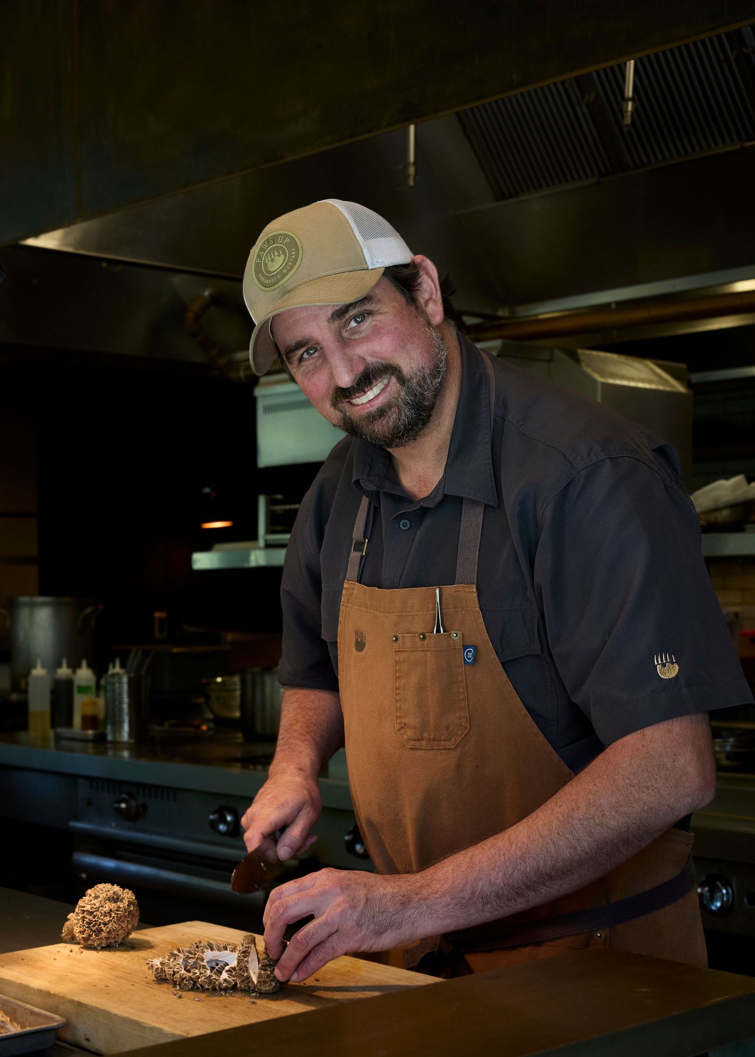 Smiling male chef in apron cutting mushrooms on wooden board in professional kitchen.