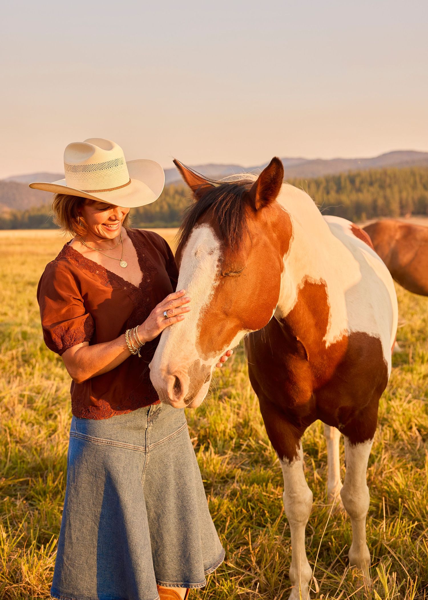 Woman in cowboy hat petting a brown and white paint horse in a golden sunset field.