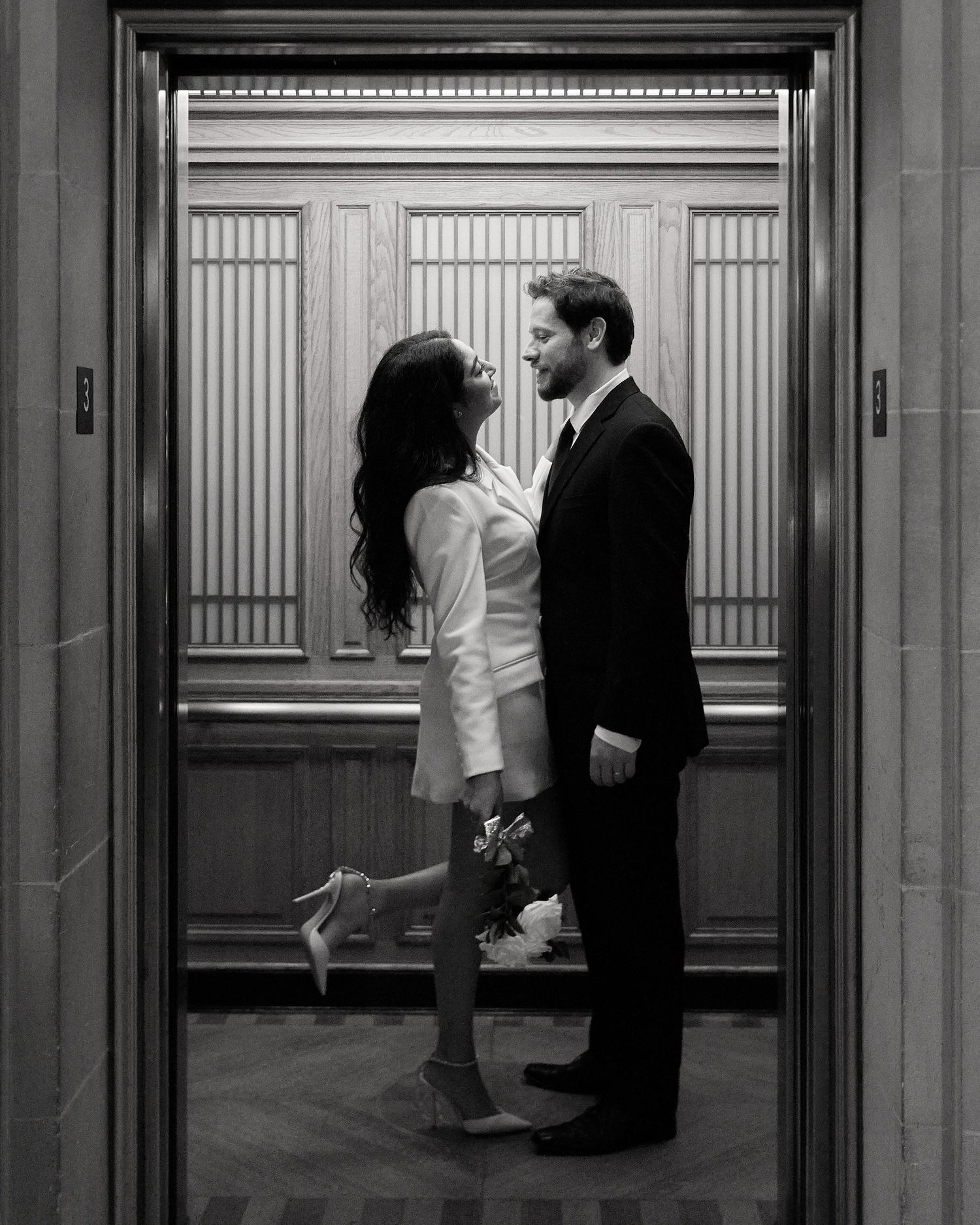 Black and white photo of couple sharing romantic moment in elevator, bride holding bouquet and heels.