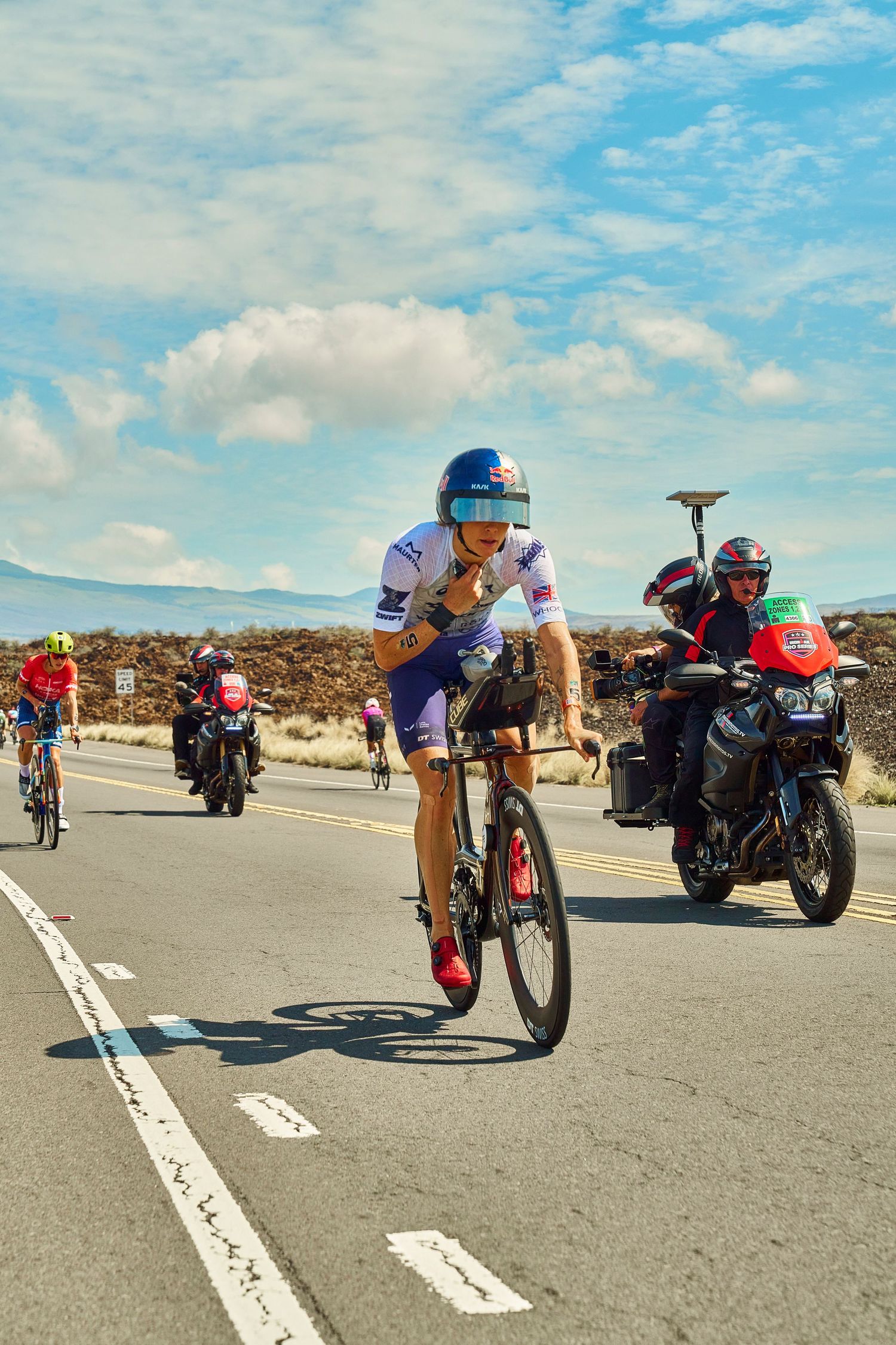 Cyclist in triathlon gear leads a race on an open road, followed by motorcycles under a partly cloudy sky.