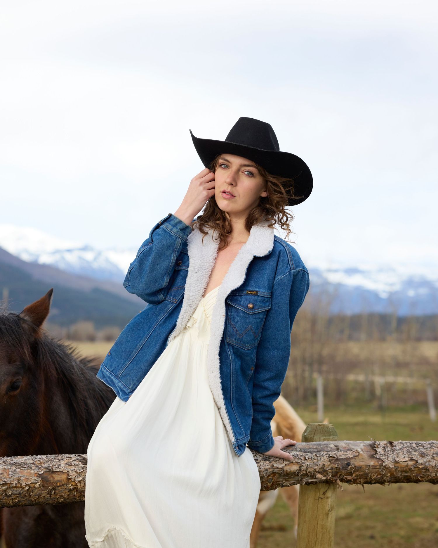 Woman in black cowboy hat and denim jacket posing on rustic fence with mountain view.