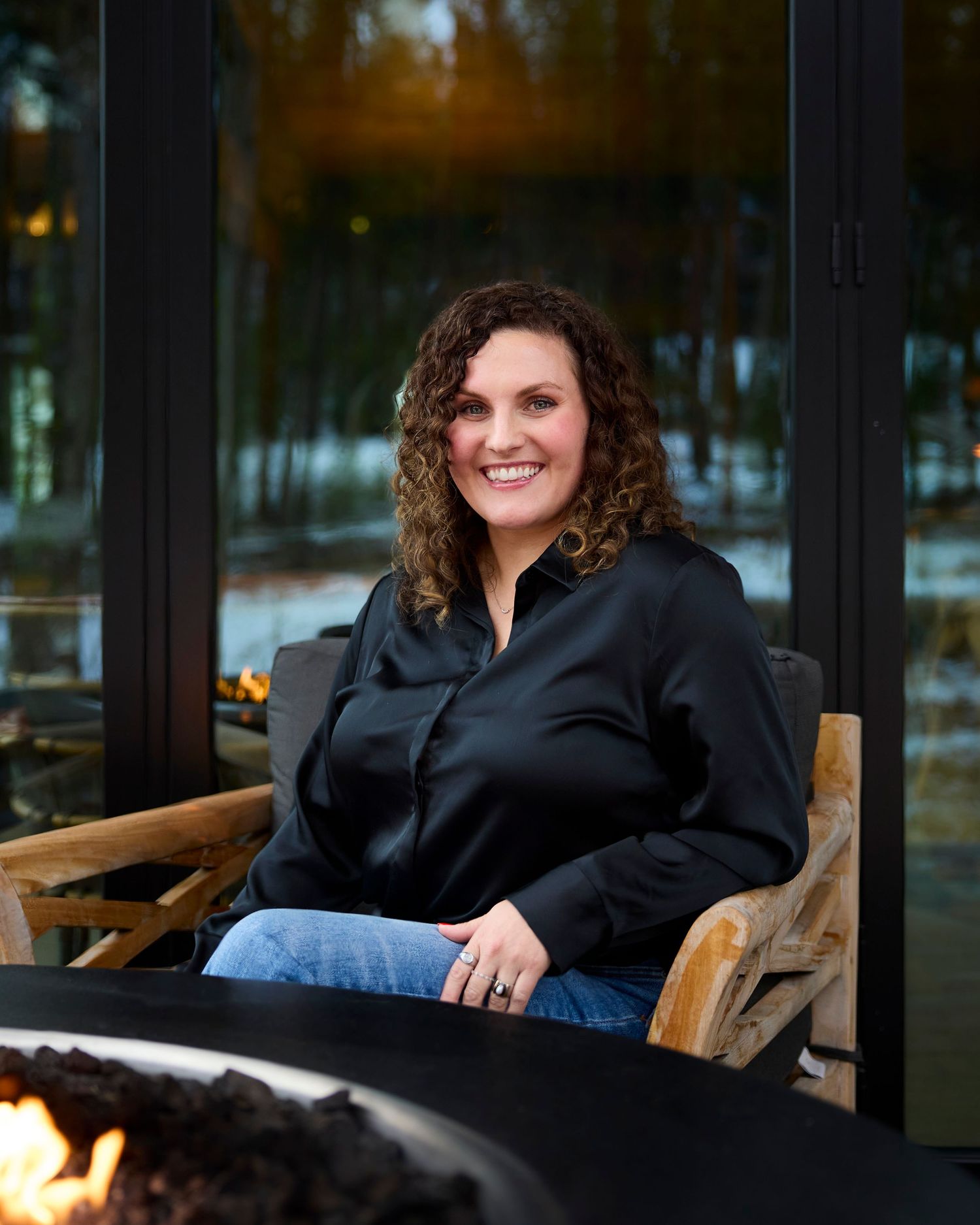 Smiling woman with curly hair in black satin blouse seated by outdoor fire pit at modern lodge.