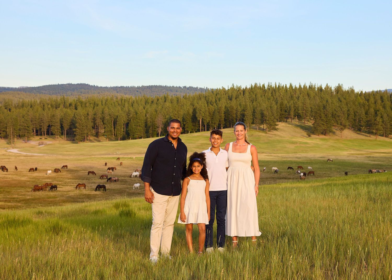 Family of four poses in a lush green meadow with grazing horses and pine forest backdrop at golden hour.