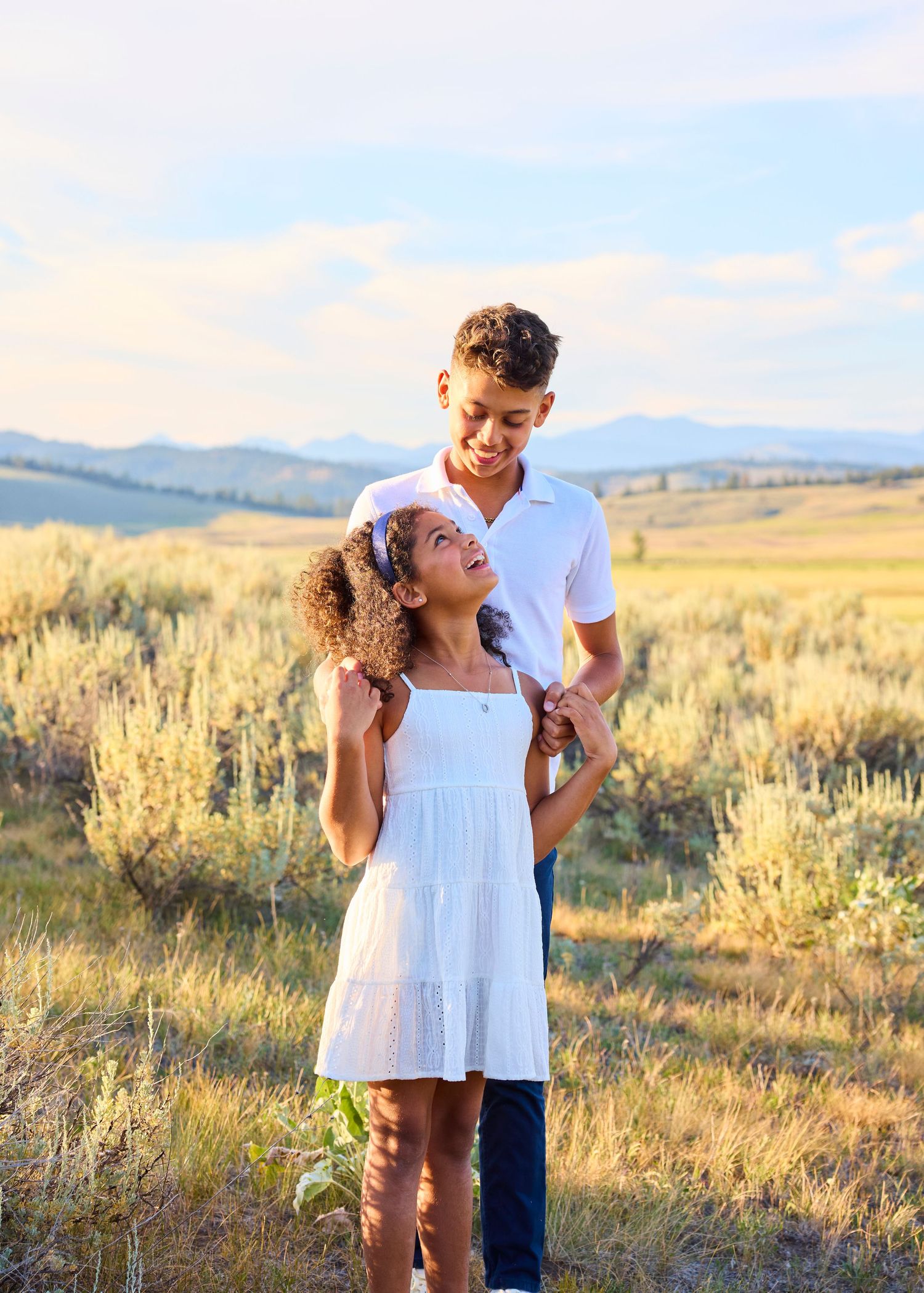 A smiling teenage boy and younger girl in white share a joyful moment in a golden sagebrush landscape.