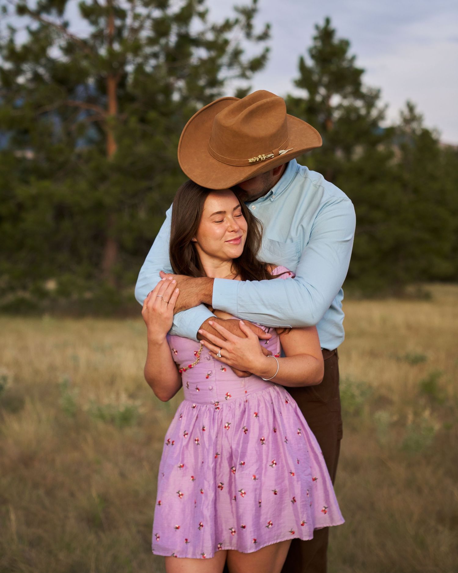 A couple embraces in a golden meadow, man in cowboy hat and blue shirt hugging woman in lavender floral dress.