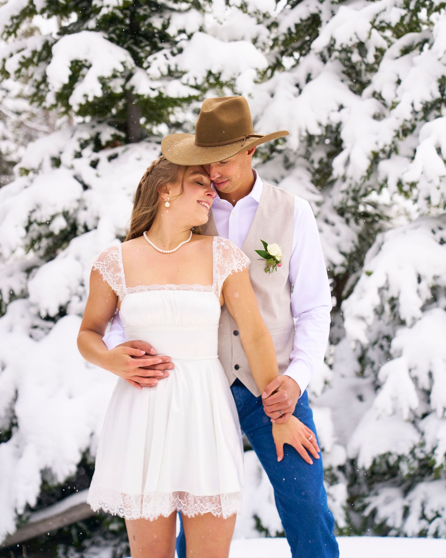 Bride and groom embrace in snowy winter forest, she wears white lace mini dress, he wears cowboy hat and vest.