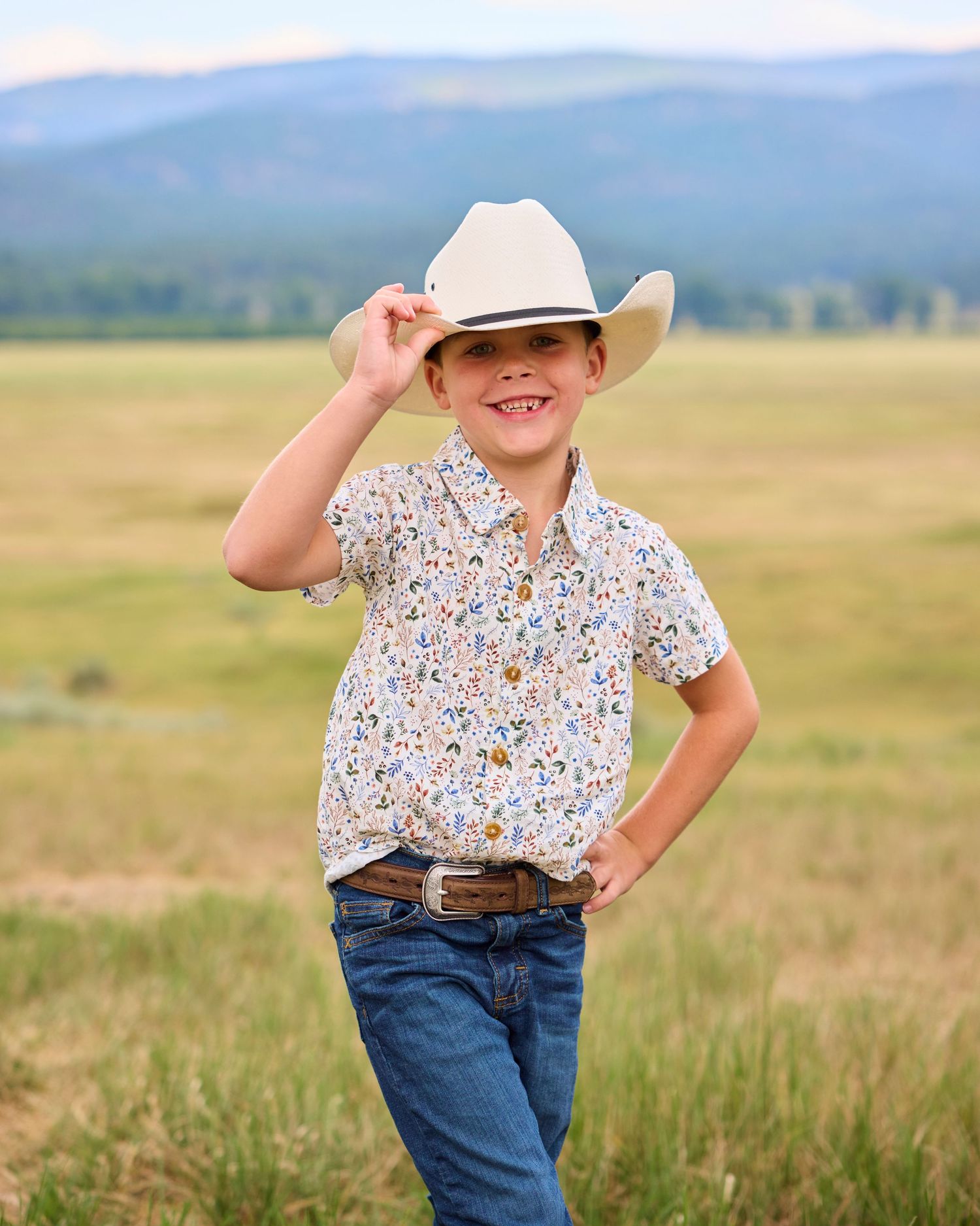 Smiling boy in white cowboy hat and floral shirt posing in green meadow with mountain backdrop.