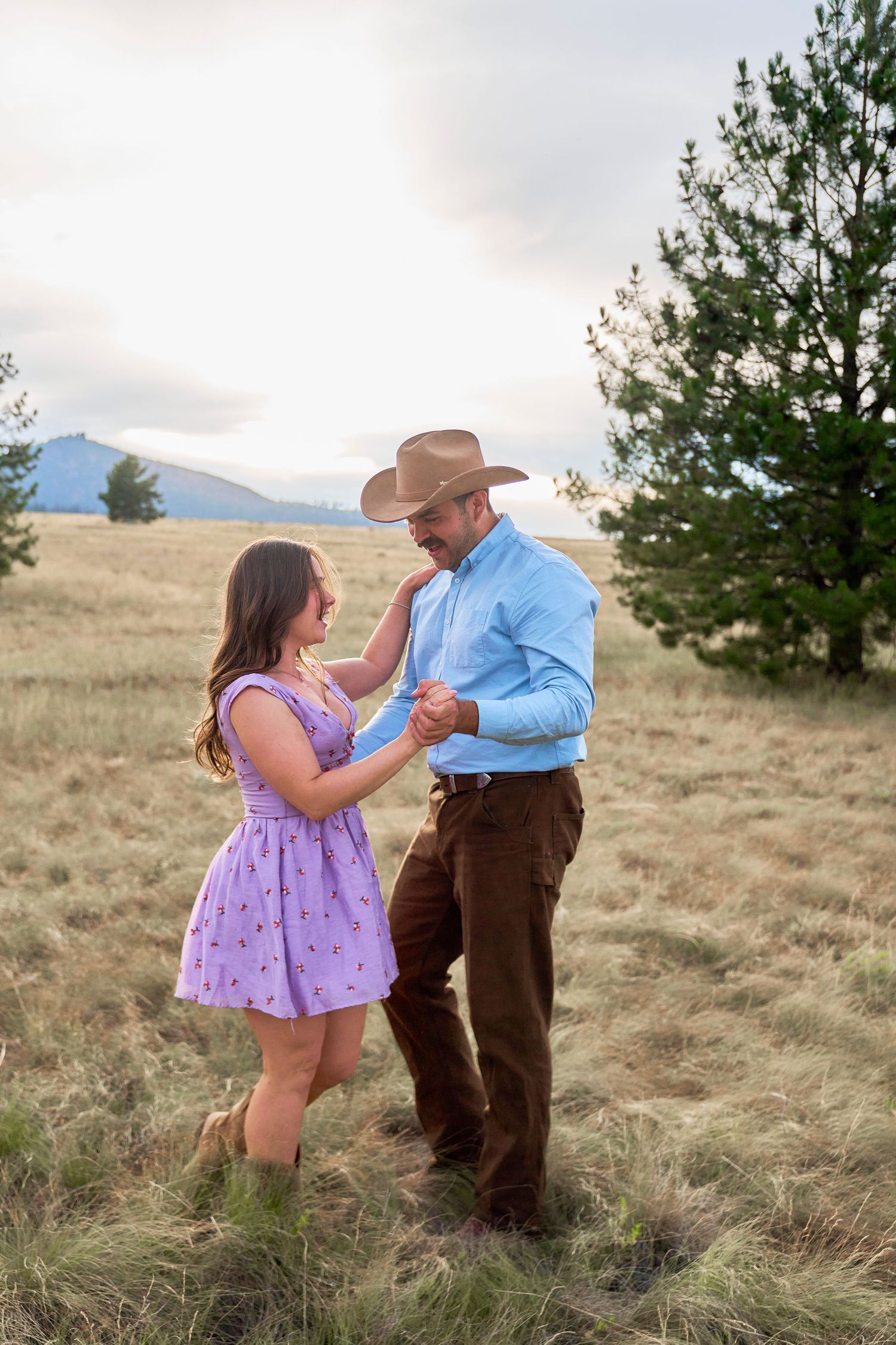 A couple dances together in a golden meadow, man wearing a cowboy hat and blue shirt, woman in purple dress.
