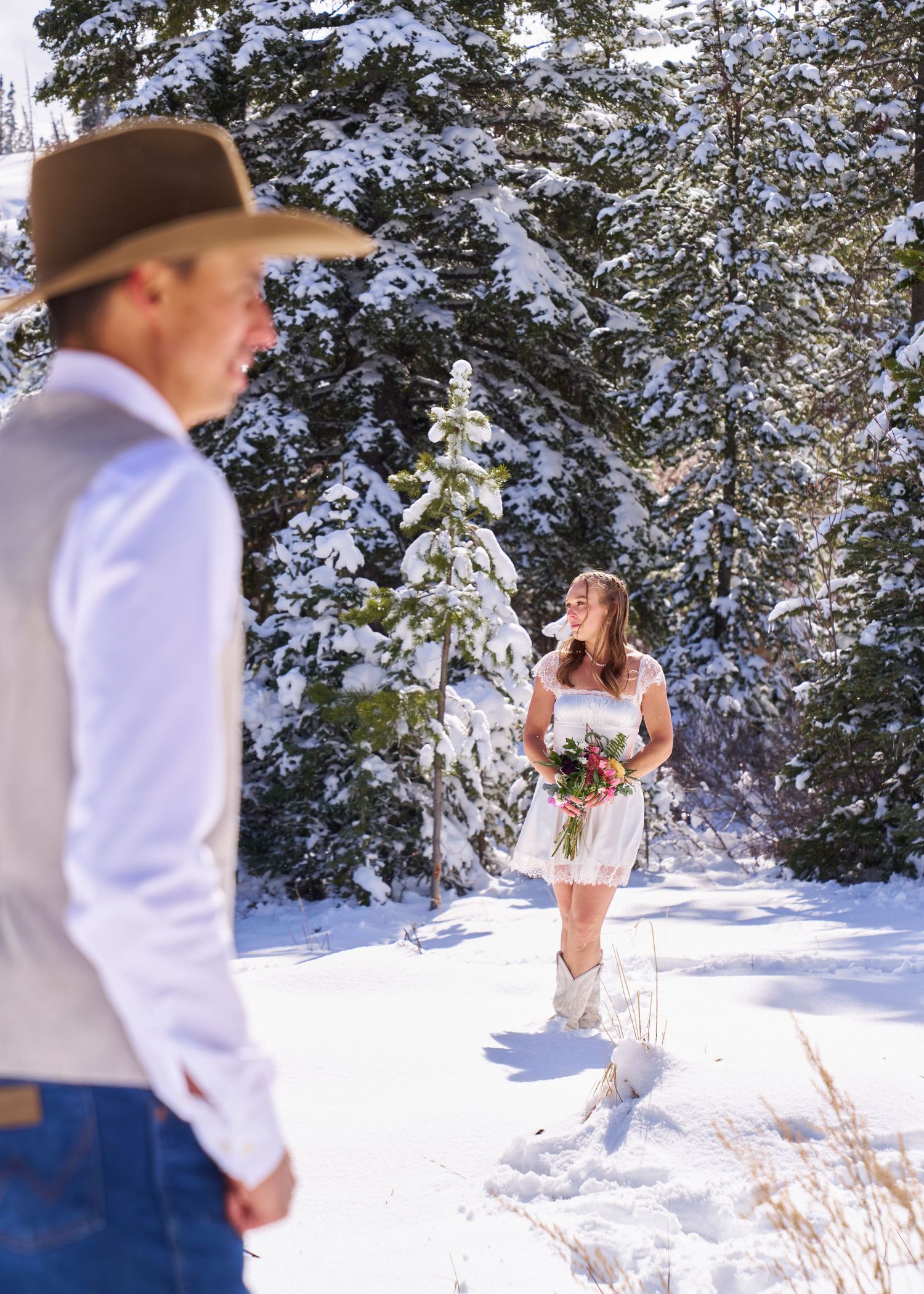 Bride in white dress and cowboy boots holds bouquet as groom watches in snowy forest setting.
