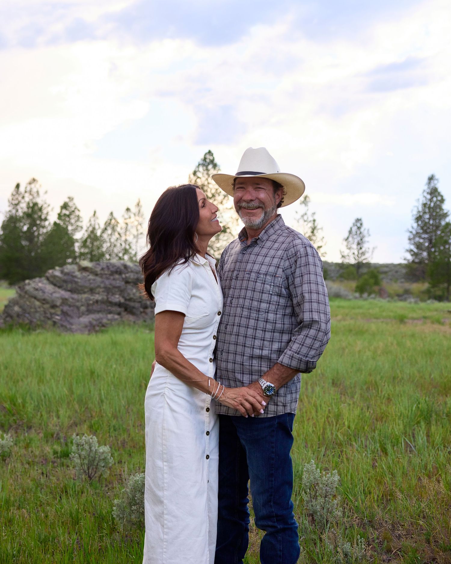 Couple embracing in a green meadow, man wearing cowboy hat and plaid shirt, woman in white dress.