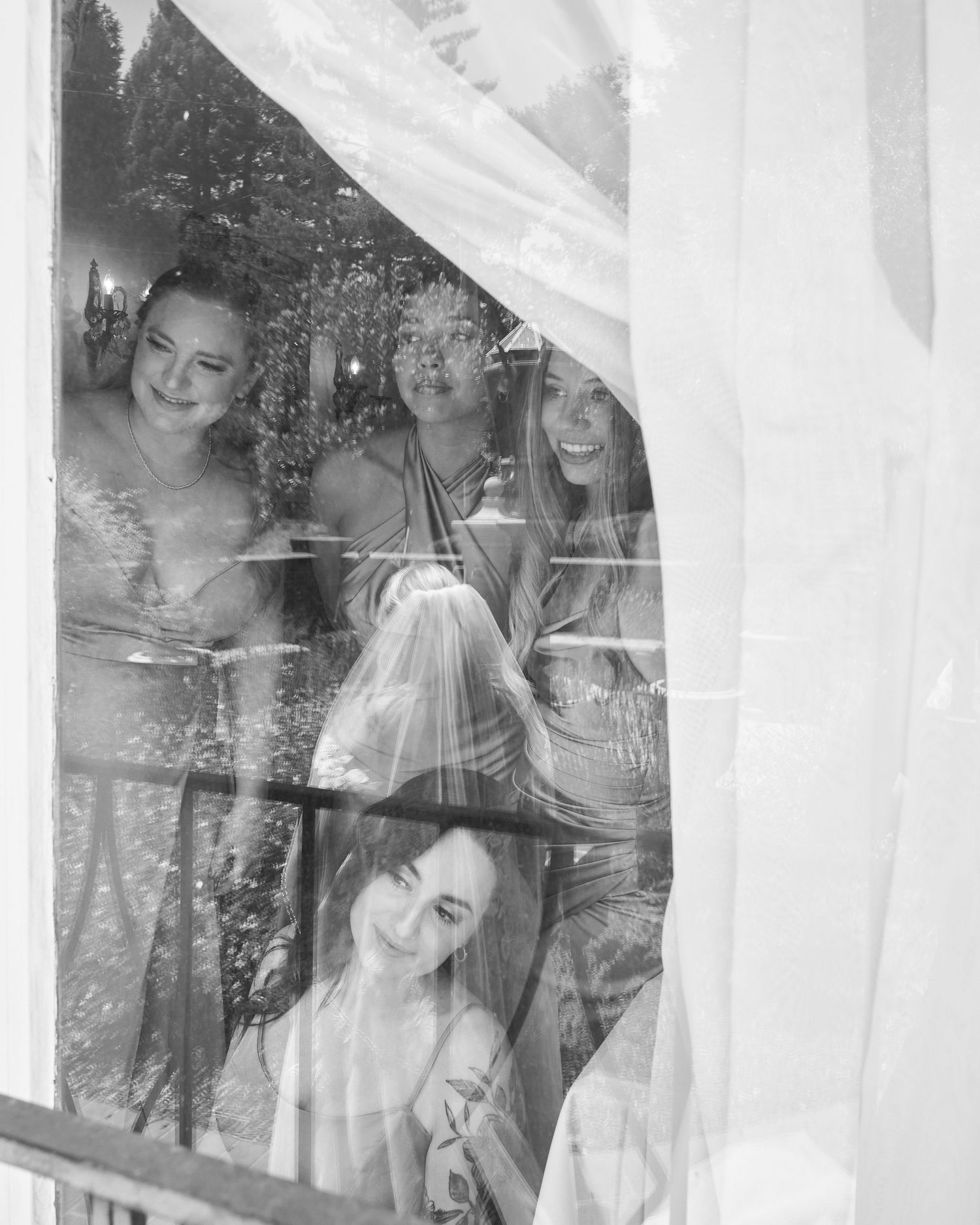 Black and white photo of smiling bride and bridesmaids peeking through sheer curtained window.