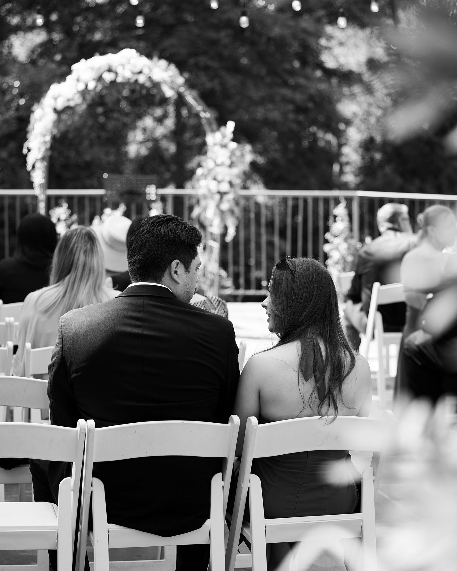 Black and white photo of wedding guests seated in white chairs before floral ceremony arch.