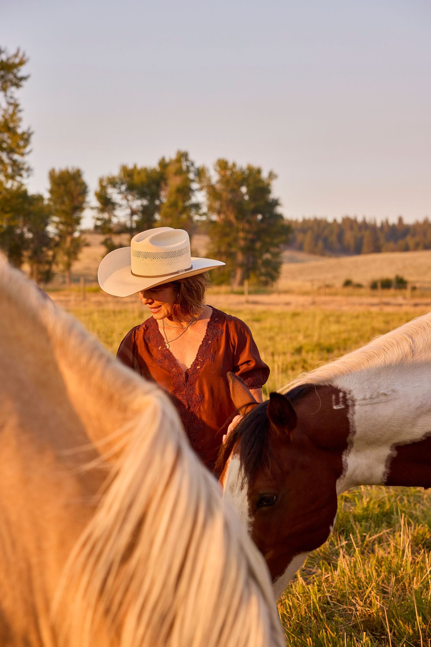 Woman in cowboy hat smiling while tending to horses in a sunlit green pasture.
