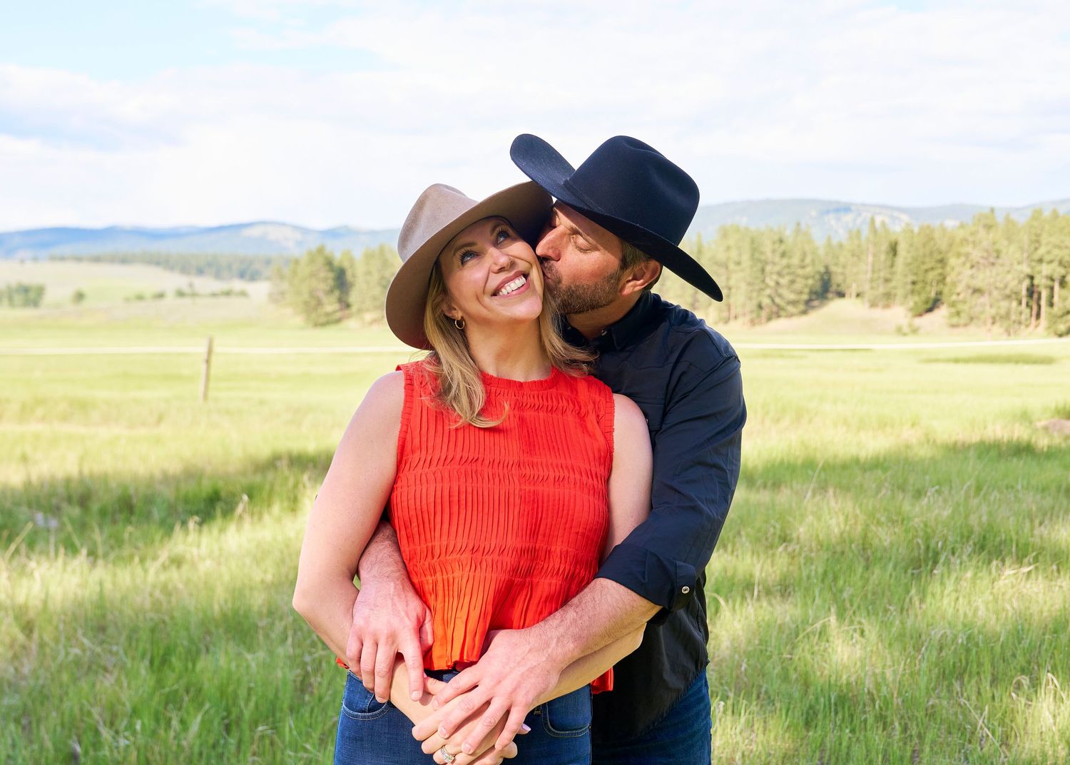 Couple embracing and smiling in a green field, man in black cowboy hat kisses woman in red top.