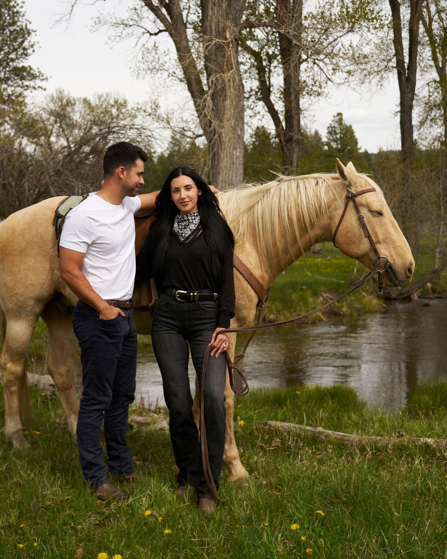 Couple standing together with palomino horse by a stream.