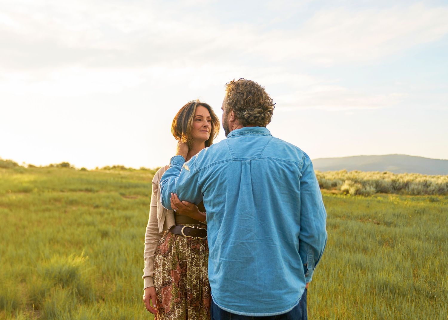 Couple standing together in a green meadow at sunset, woman in floral skirt smiling at bearded man.
