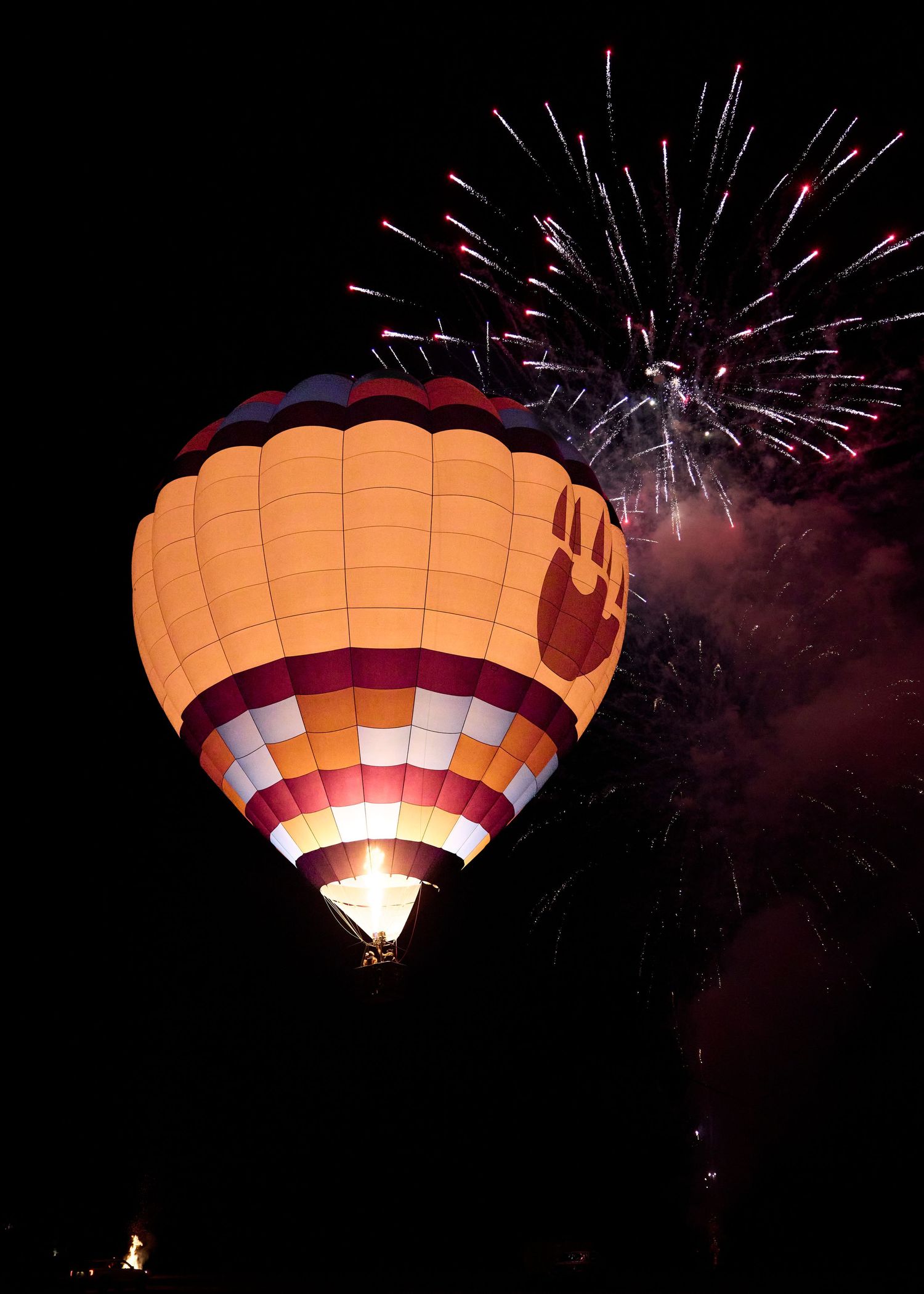 An illuminated hot air balloon glows against a dark night sky with fireworks bursting behind it.