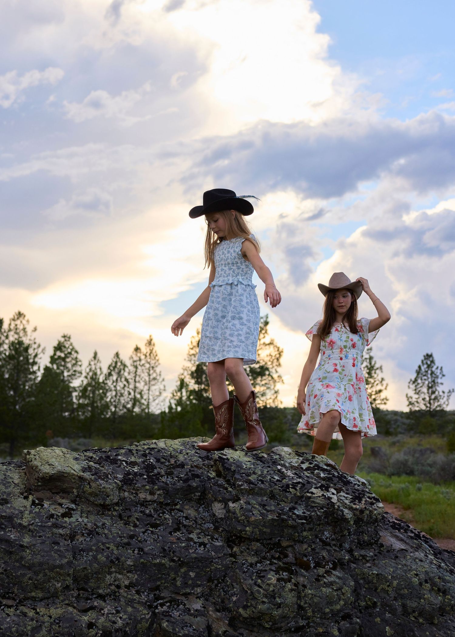 Two young girls in cowboy hats and boots standing on a rock at sunset in a rural landscape.
