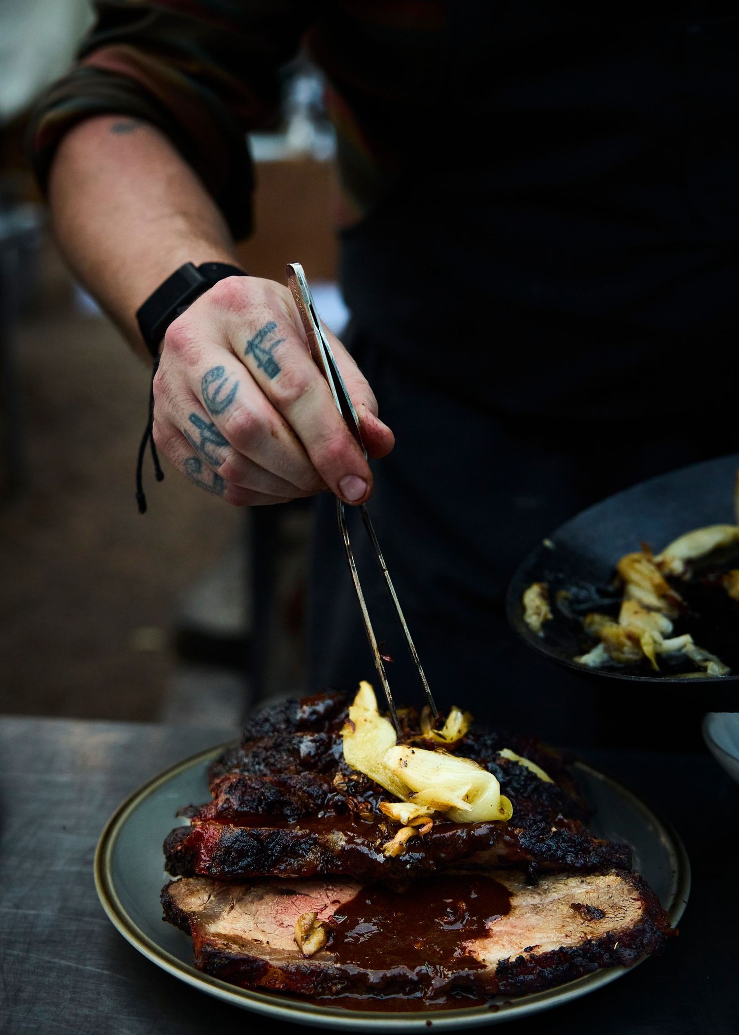Tattooed chef using tongs to plate glazed BBQ brisket on ceramic dish in kitchen.