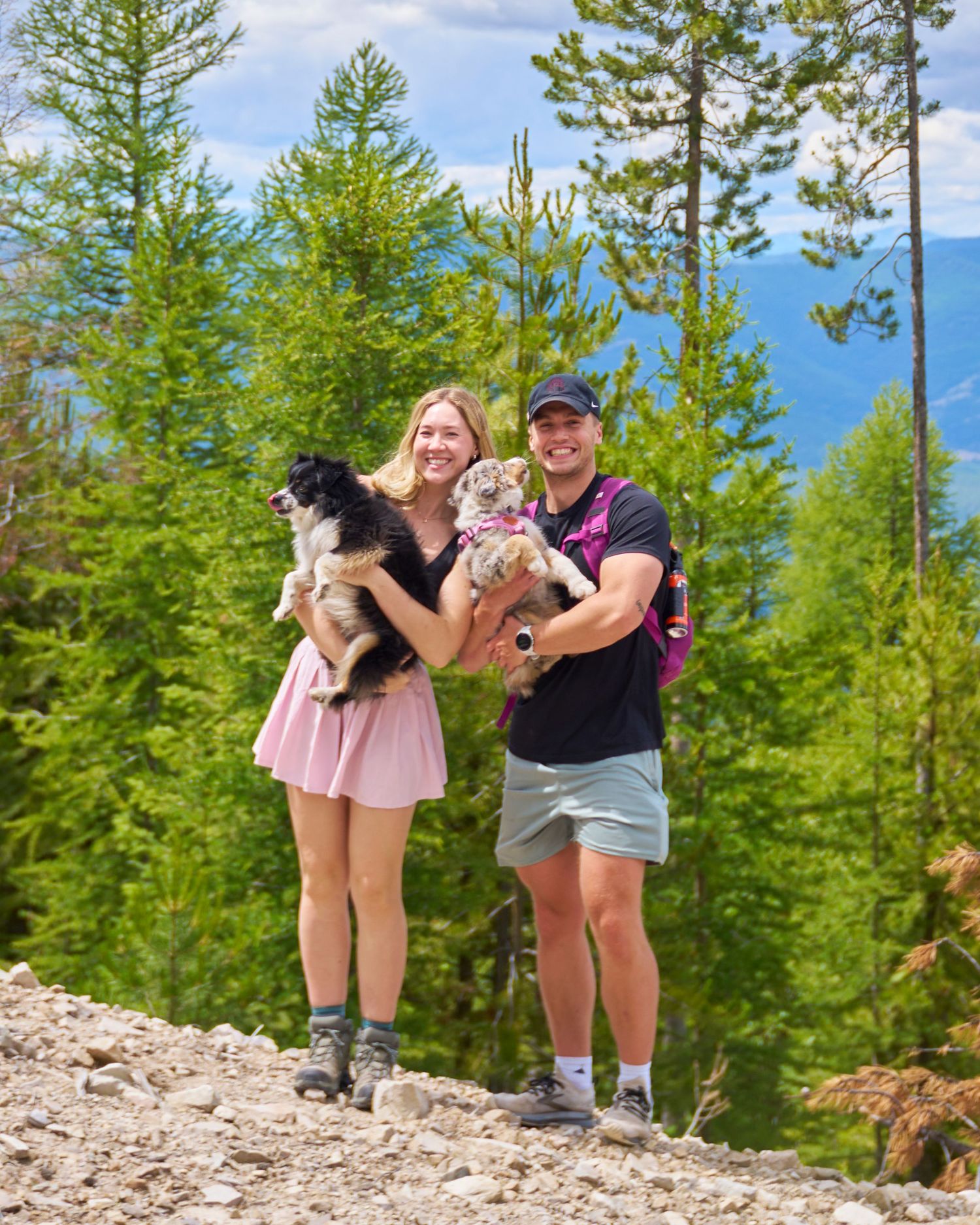 Smiling couple holds two small dogs on a rocky mountain trail surrounded by green pine trees.