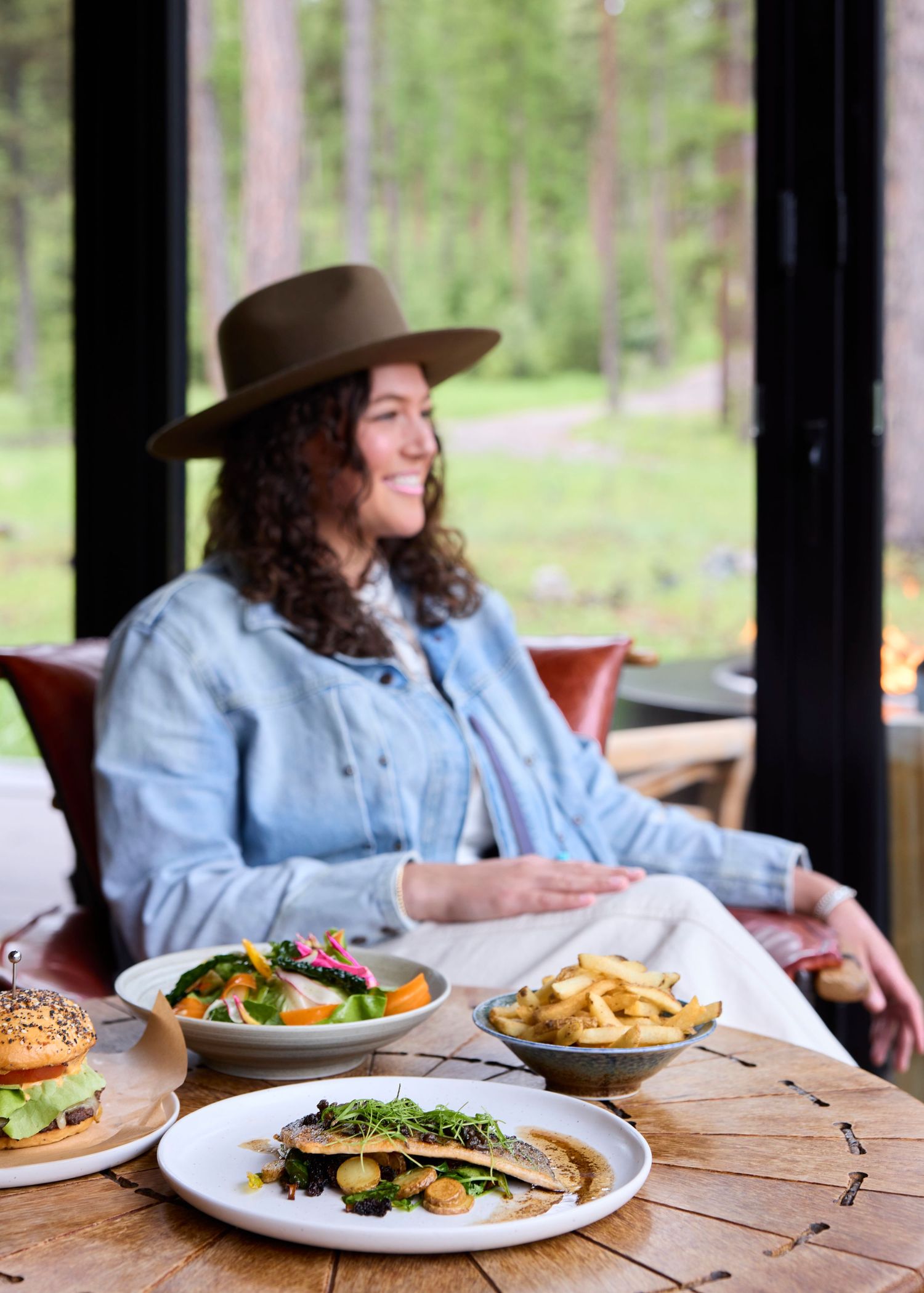 Smiling woman in wide-brim hat seated at table with gourmet dishes including fish fillet.
