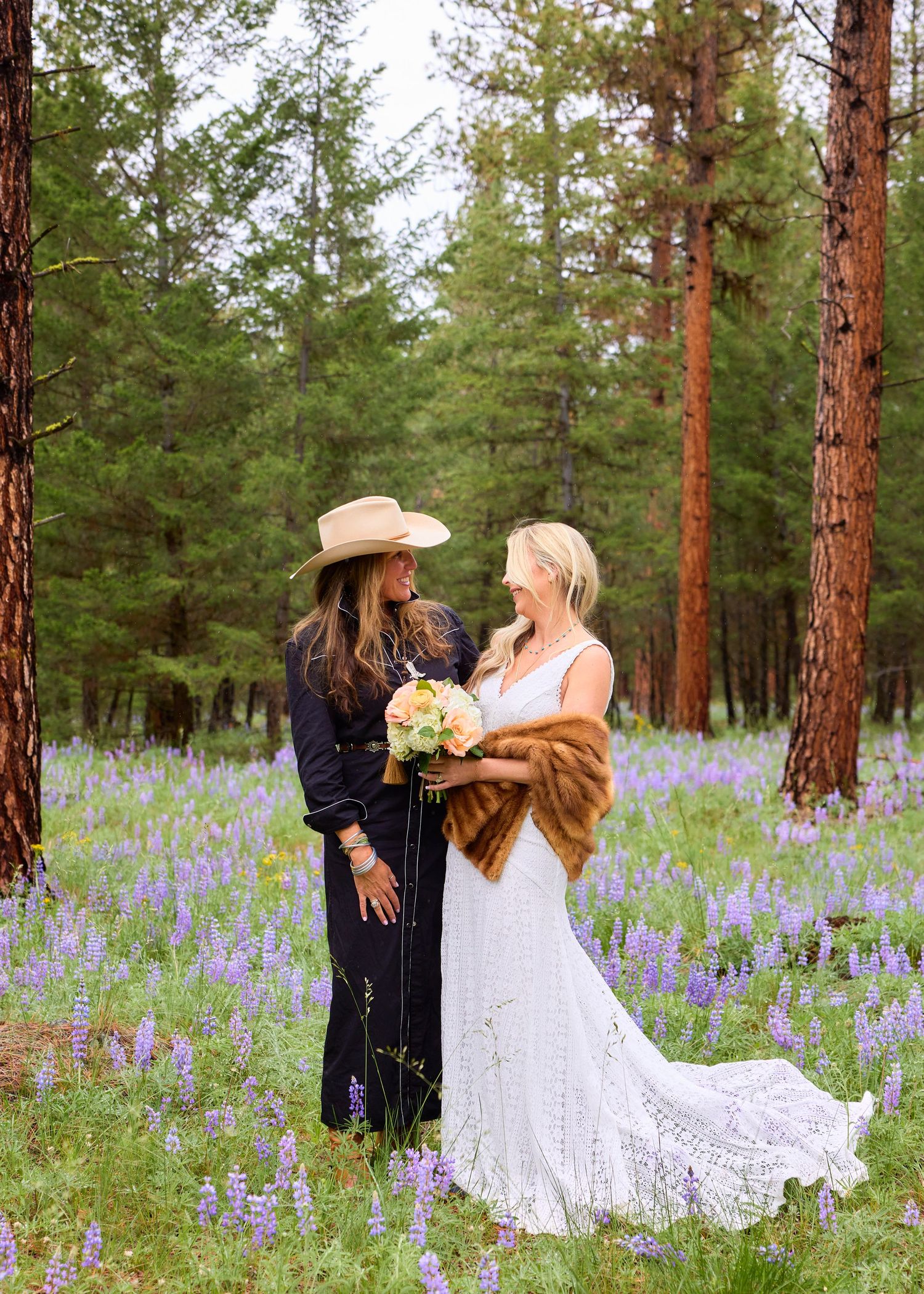 Officiant in wide-brim hat faces bride in white gown with fur wrap, surrounded by purple lupines in pine forest.