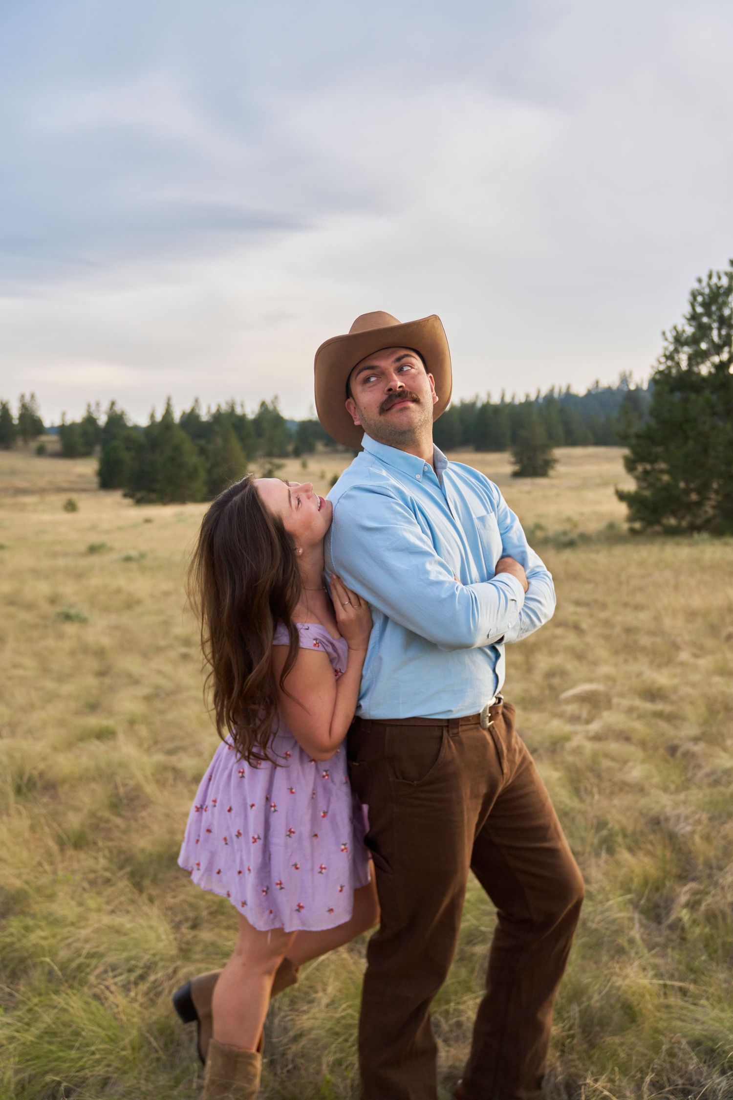 Man in cowboy hat with arms crossed stands in field as woman in purple dress leans against his back playfully.