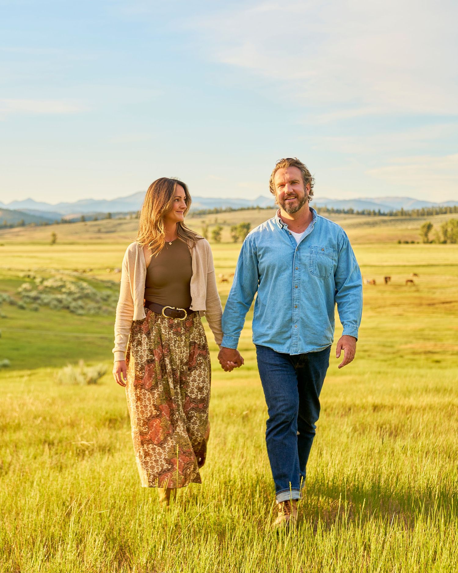 Couple walking hand in hand through a bright green meadow with mountains and grazing animals in the background.
