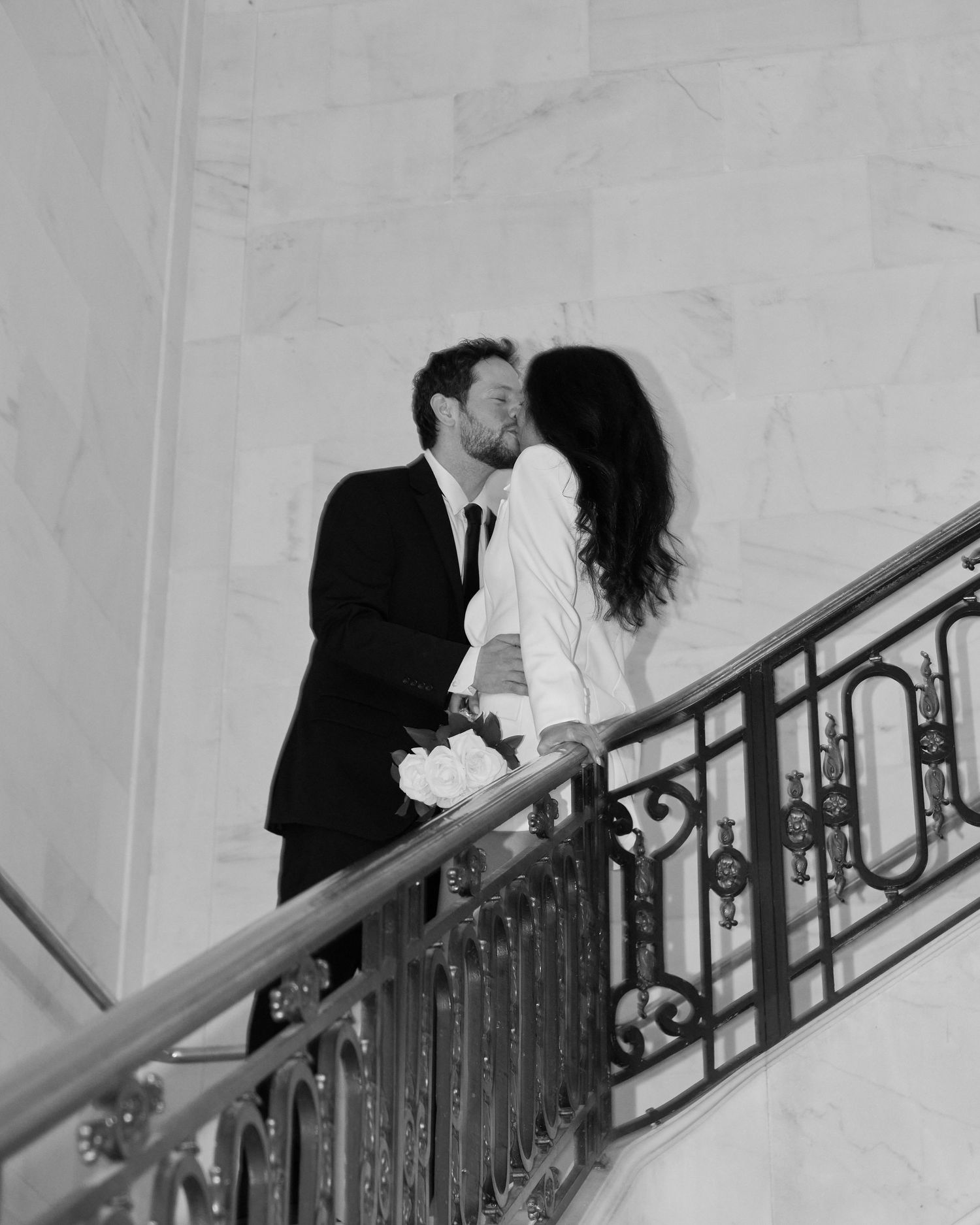 Black and white wedding photo of couple kissing on an ornate staircase railing, groom holding white roses bouquet.